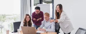 Foto von 4 jungen Mitarbeitern und Mitarbeiterinnen die lachend im Besprechungsraum vor einem Laptop sitzen bzw. auch stehen.