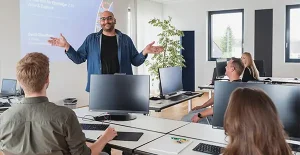 Lächelnder IT Trainer unterrichtet Schulungsteilnehmer im Schulungsraum. Hinter ihm wird eine Powerpoint Präsentation an die Wand gestrahlt.